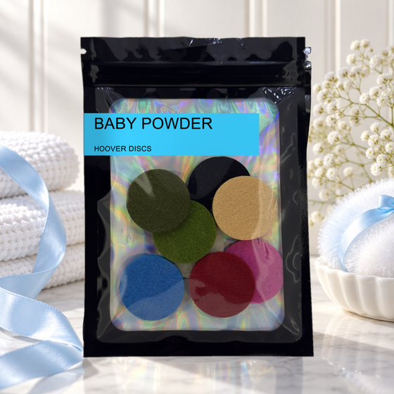 Pack of 8 Baby Powder scented hoover discs in its sealed black foil packet beside folded white towels, blue ribbon and gypsophila
