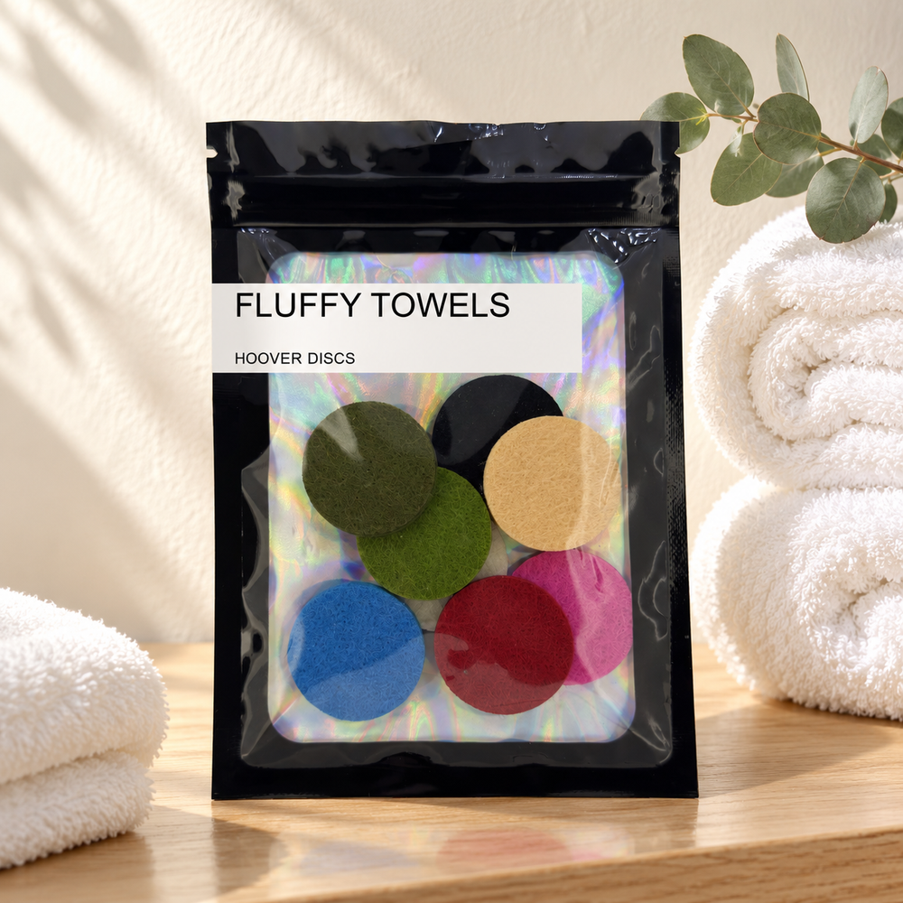 Pack of 8 Fluffy Towels scented hoover discs in its sealed black foil packet beside rolled white towels and a sprig of eucalyptus