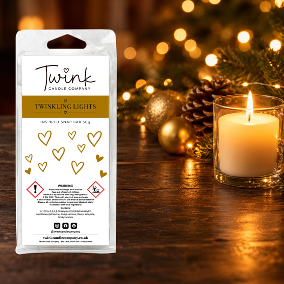 Twinkling Lights inspired soy wax melt snap bar by Twink Candle Company, stood on a dark wood table beside a lit candle, gold baubles and a pine cone