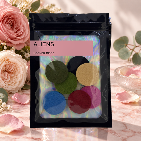 Pack of 8 Aliens scented hoover discs in its sealed black foil packet beside pink roses and eucalyptus on pale marble