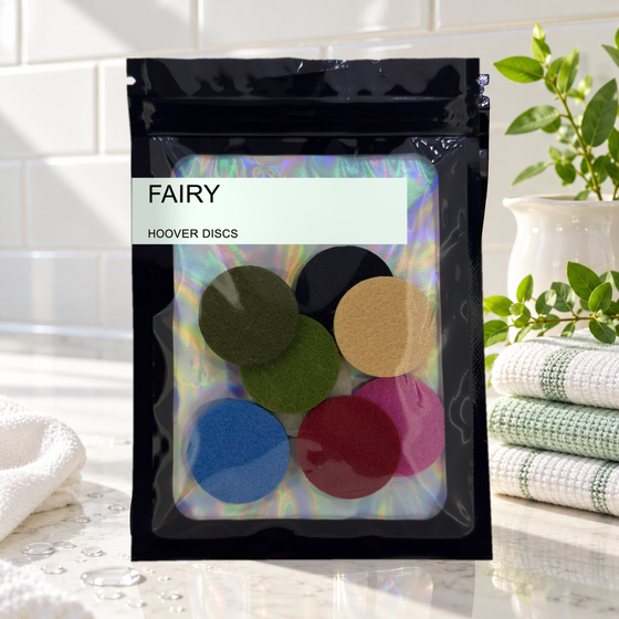 Pack of 8 Fairy scented hoover discs in its sealed black foil packet beside folded white and green towels and a leafy plant