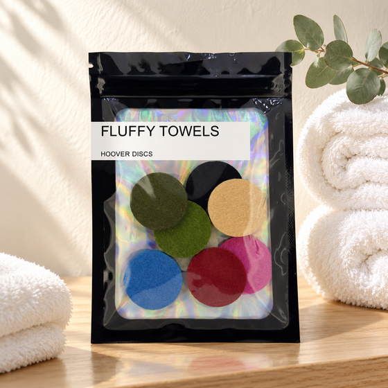 Pack of 8 Fluffy Towels scented hoover discs in its sealed black foil packet beside rolled white towels and a sprig of eucalyptus